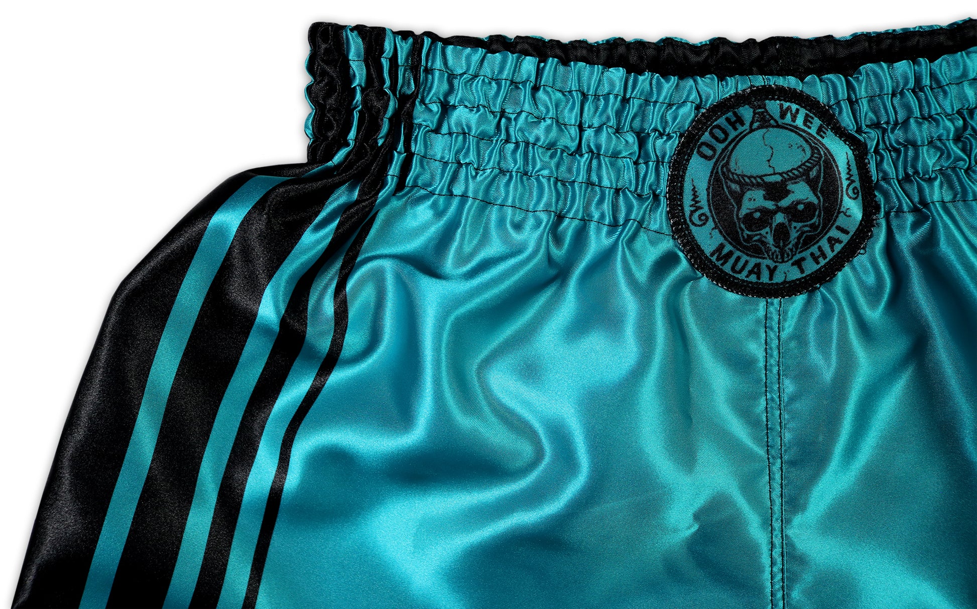 Close-up of turquoise Muay Thai shorts featuring black side stripes, an elastic waistband, and a round 'OOH-WEE Muay Thai' logo with a skull on the front.